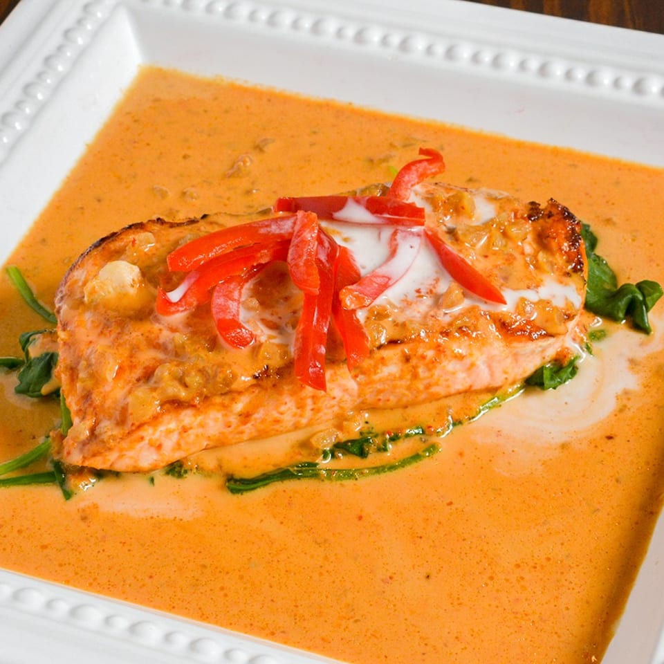 Salmon fillet with red pepper strips, cream sauce, and spinach on a white rectangular plate.