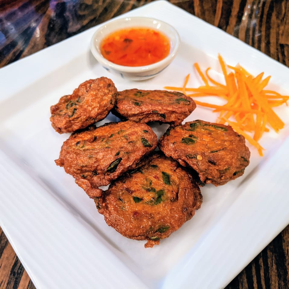 Five golden-brown Thai fish cakes with sweet chili sauce and shredded carrots on a white plate.