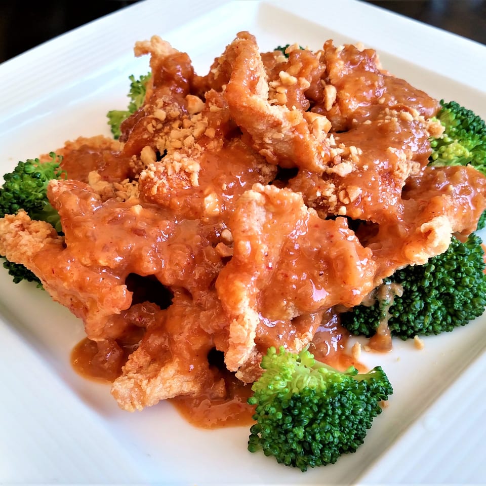Crispy chicken with peanut sauce and chopped peanuts, served over steamed broccoli on a white plate.