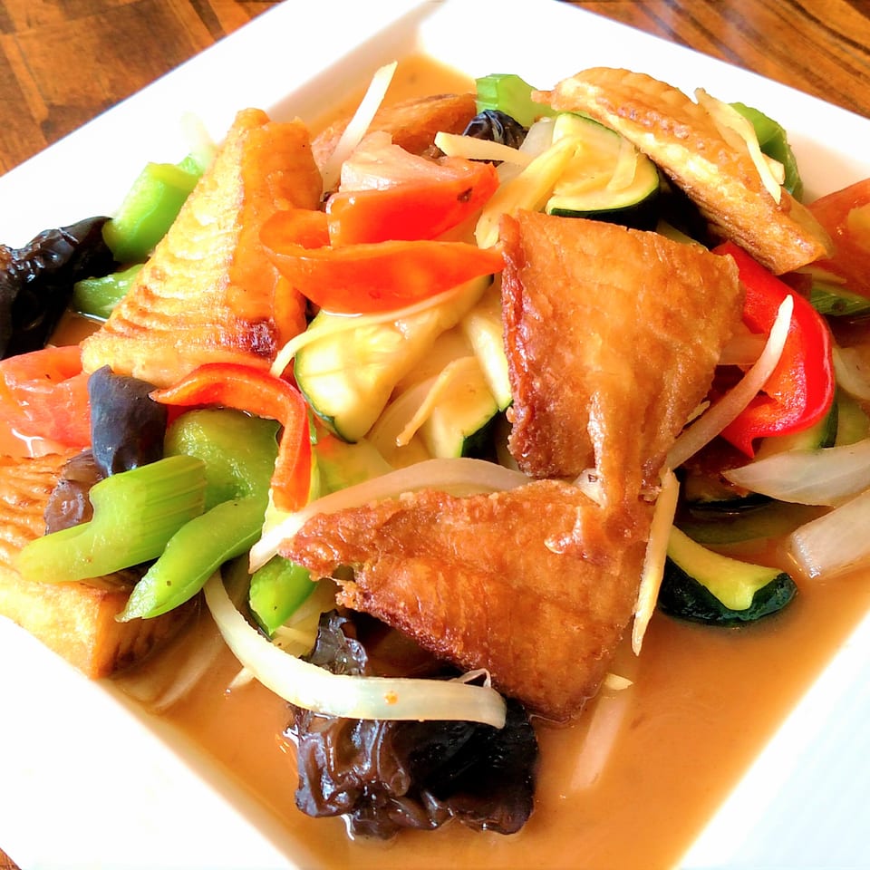 Crispy fried fish with colorful vegetables and mushrooms in a savory sauce on a white plate.